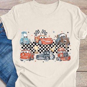 May include: A beige t-shirt features a vintage-style illustration of various cartoon cars, including a red race car, a firetruck, a police car, and a tow truck. The cars are arranged against a black and white checkered background, with stars and checkered flags adding to the design. The overall theme is reminiscent of classic racing imagery and children's cartoons.