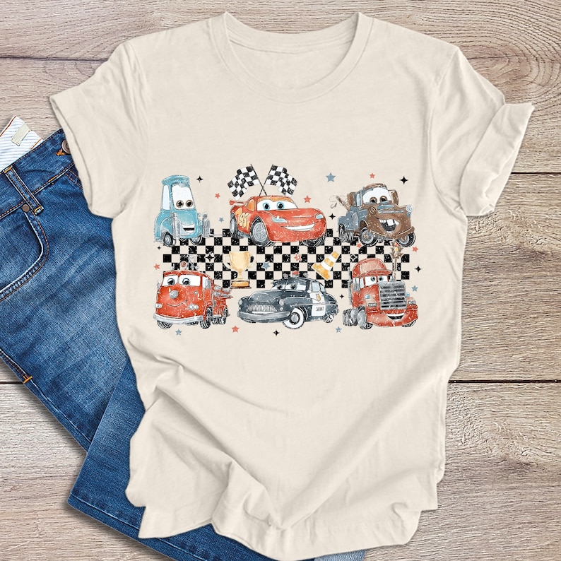May include: A beige t-shirt features a vintage-style illustration of various cartoon cars, including a red race car, a firetruck, a police car, and a tow truck. The cars are arranged against a black and white checkered background, with stars and checkered flags adding to the design. The overall theme is reminiscent of classic racing imagery and children's cartoons.