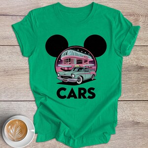 May include: Green t-shirt with a retro car design, a diner, and the word "CARS". The graphic includes a teal car, a pink diner, and black Mickey Mouse ears. The shirt is on a wooden surface.