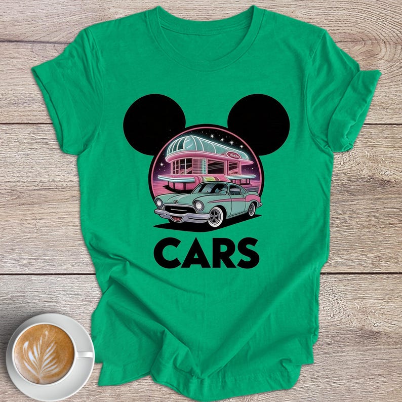 May include: Green t-shirt with a retro car design, a diner, and the word "CARS". The graphic includes a teal car, a pink diner, and black Mickey Mouse ears. The shirt is on a wooden surface.