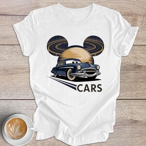 May include: White t-shirt with a cartoon car design. The design features a blue car with a smiling face, a yellow sun, and a road design. The word "CARS" is printed below the car in a bold font.