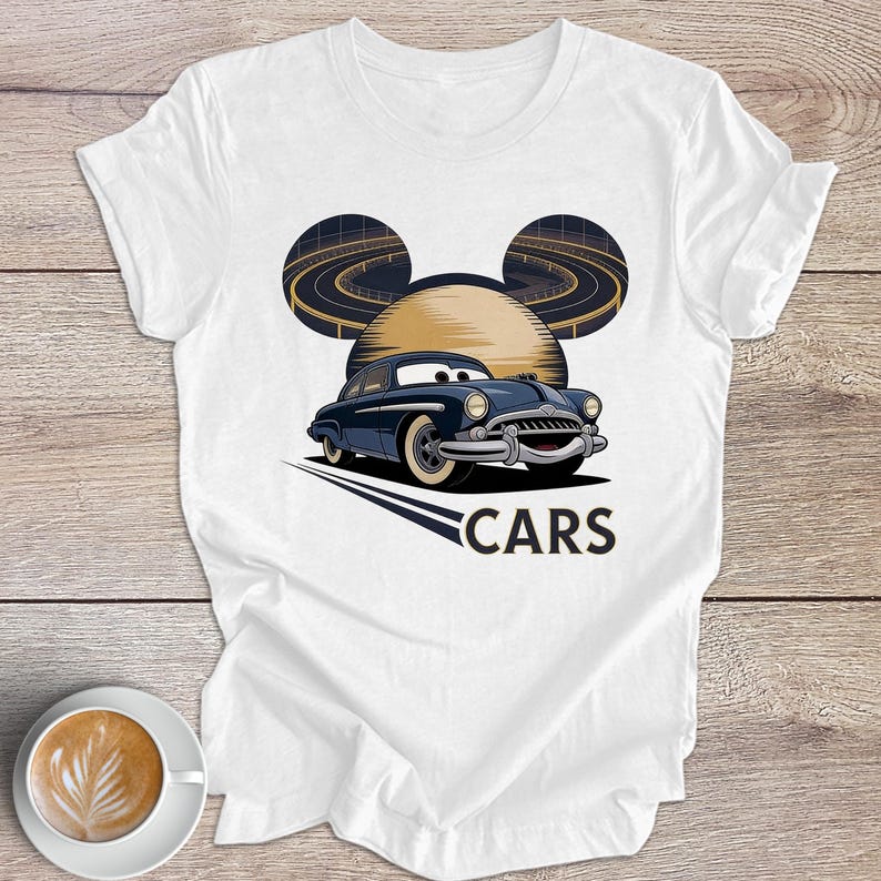 May include: White t-shirt with a cartoon car design. The design features a blue car with a smiling face, a yellow sun, and a road design. The word "CARS" is printed below the car in a bold font.