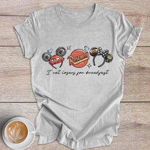 Peut inclure: Un t-shirt gris clair avec un motif graphique inspir&eacute; du film Cars. Le motif comprend des images de Flash McQueen, un logo "Rusteze" et un logo "95". Le texte "I eat losers for breakfast" est imprim&eacute; sous les graphiques.