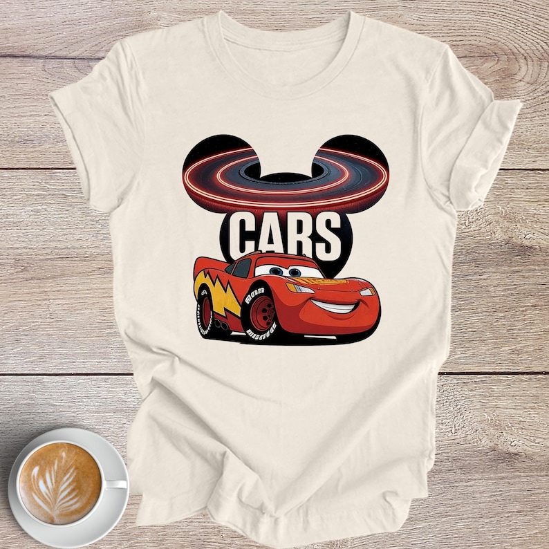 May include: Cream-coloured t-shirt with a graphic of Lightning McQueen from the film Cars. The design includes the word "CARS", a race track, and the character's smiling face. The shirt is on a wooden surface.
