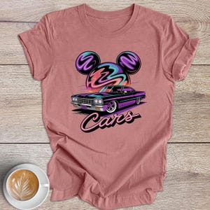 May include: A dusty rose t-shirt with a graphic of a classic car, a colourful sunset, and Mickey Mouse ears. The word "Cars" is written in a script font below the car. A cup of coffee sits in the lower left corner.