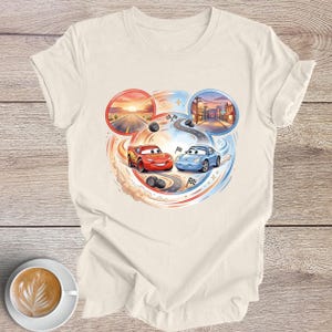 Peut inclure: T-shirt cr&egrave;me avec un motif color&eacute; de deux voitures de course de dessins anim&eacute;s, Flash McQueen et Sally Carrera, sur une piste. Le motif comprend un coucher de soleil et une sc&egrave;ne de ville, avec des drapeaux de course et des traces de pneus.