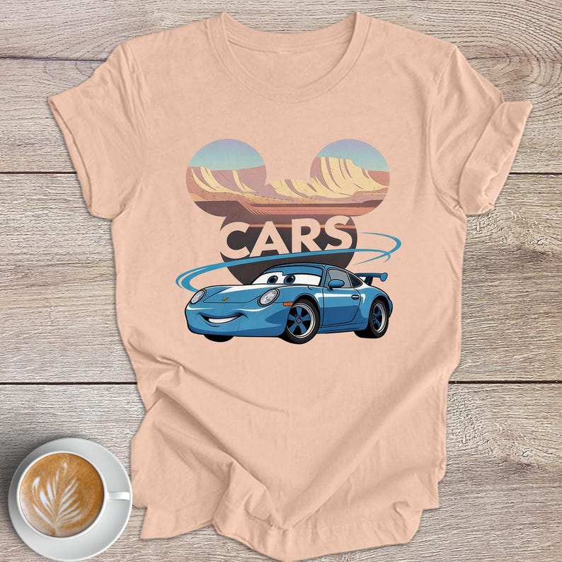 May include: Pale pink t-shirt with a cartoon car design. The blue car has a smiling face and is set against a desert landscape with the word "CARS". The design includes a stylized logo above the car.