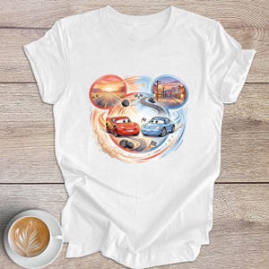 Peut inclure: T-shirt blanc avec un motif color&eacute; de deux voitures de course de dessins anim&eacute;s, une rouge et une bleue, sur un circuit. Le design comprend des sc&egrave;nes de coucher de soleil et de ville dans des cadres circulaires, avec des traces de pneus et des drapeaux &agrave; damier.