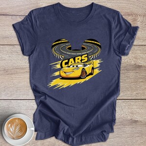 May include: Navy blue t-shirt with a yellow cartoon car graphic, the word "CARS", and a race track design. The shirt is made of a soft material, suitable for everyday wear.