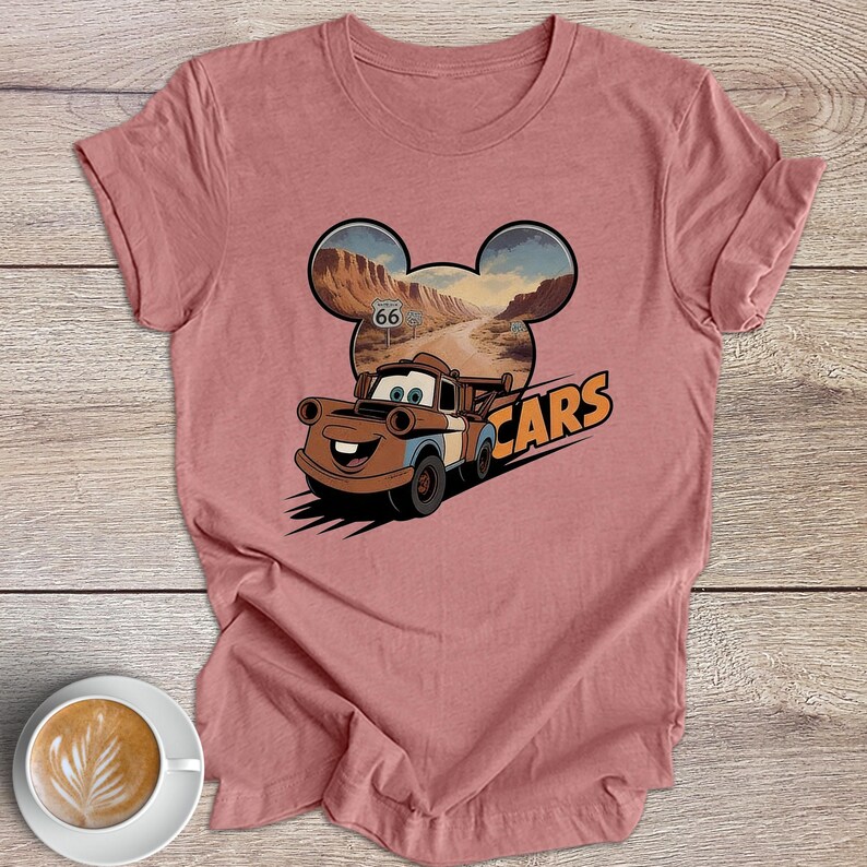 May include: A dusky pink t-shirt with a graphic of the character Mater from the Disney Pixar film "Cars." The design includes a Route 66 sign and the word "CARS" in orange. The shirt is displayed on a wooden surface.