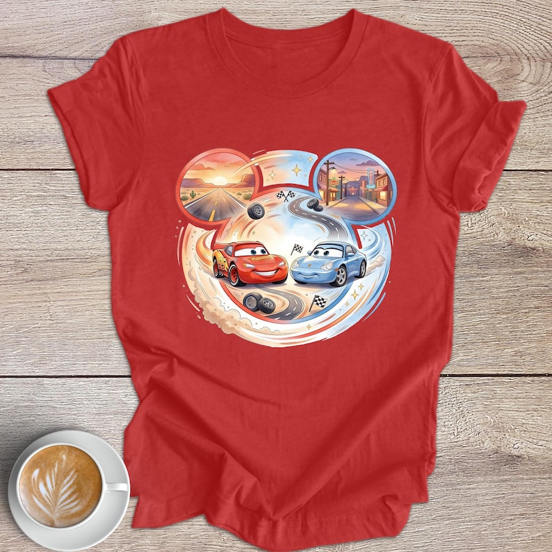 Peut inclure: T-shirt rouge avec un motif de dessin anim&eacute; de deux voitures de course, une rouge et une bleue, sur une piste. Le motif comprend un coucher de soleil dans le d&eacute;sert et une sc&egrave;ne de ville, avec des traces de pneus et des drapeaux &agrave; damier. Une tasse de caf&eacute; est en bas &agrave; gauche.