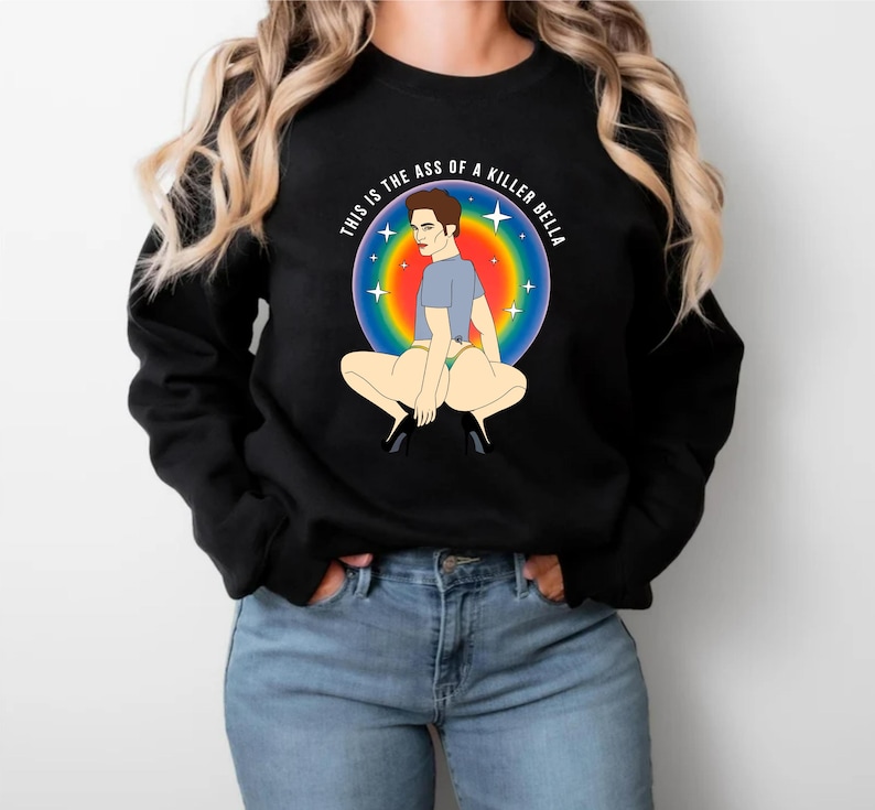 Peut inclure: Sweat-shirt noir avec un dessin color&eacute; d'une personne accroupie, entour&eacute;e d'un cercle arc-en-ciel et du texte "THIS IS THE ASS OF A KILLER BELLA". L'illustration est de style cartoon.