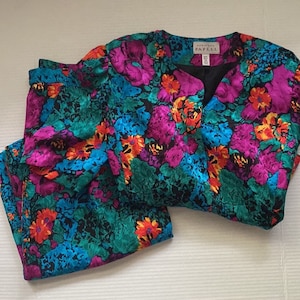 Puede incluir: Un conjunto de falda y chaqueta de estampado floral en tonos rosa, azul, verde y amarillo. La chaqueta tiene cuello en V y la etiqueta "Adrianna Papell" es visible en la parte delantera.