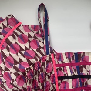May include: A pink and brown patterned fabric with a pink and blue geometric design. The fabric has a pink and blue trim and a blue strap.