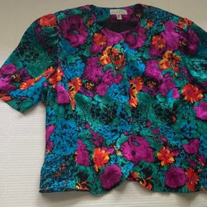 Puede incluir: Una blusa de manga corta con estampado floral y cuello en V. La blusa est&aacute; hecha de una tela sedosa y presenta una mezcla vibrante de colores, incluyendo turquesa, morado, naranja y amarillo.