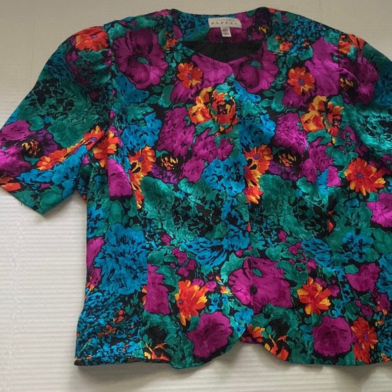 Puede incluir: Una blusa de manga corta con estampado floral y cuello en V. La blusa est&aacute; hecha de una tela sedosa y presenta una mezcla vibrante de colores, incluyendo turquesa, morado, naranja y amarillo.