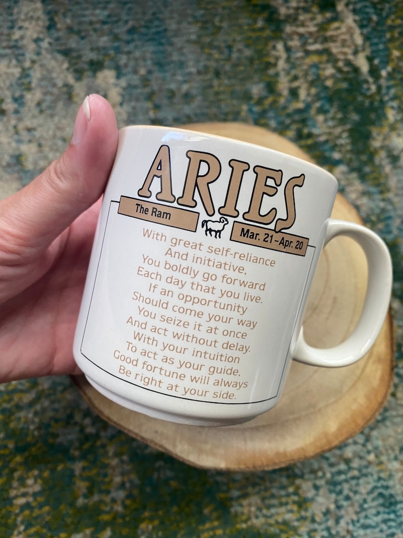 Może przedstawiać: Biały ceramiczny kubek z brązowo-złotym wzorem przedstawiającym znak zodiaku Baran. Kubek ma uchwyt i napis "The Ram" oraz "Mar. 21-Apr. 20". Tekst brzmi: "Z wielką pewnością siebie i inicjatywą. Odważnie idziesz naprz&oacute;d każdego dnia swojego życia. Jeśli nadarzy się okazja, chwytasz ją natychmiast i działasz bez zwłoki. Z intuicją jako przewodnikiem. Szczęście zawsze będzie po twojej stronie."