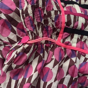 May include: A white cotton dress with a pink and purple geometric pattern. The dress has a pink elastic band at the waist with a pink button.