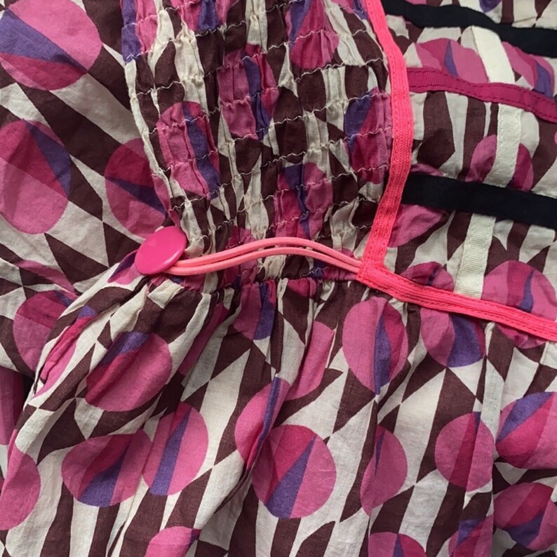 May include: A white cotton dress with a pink and purple geometric pattern. The dress has a pink elastic band at the waist with a pink button.