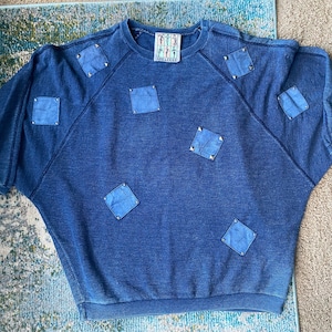 May include: A blue denim sweatshirt with a relaxed fit and a crew neck. The sweatshirt has several square patches sewn onto the front and shoulders. The patches are a lighter shade of blue than the sweatshirt.