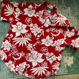 May include: A red short-sleeved button-up shirt with a white floral pattern. The shirt has a collar and a button closure. The brand name "Hawaiian" is visible on the collar.
