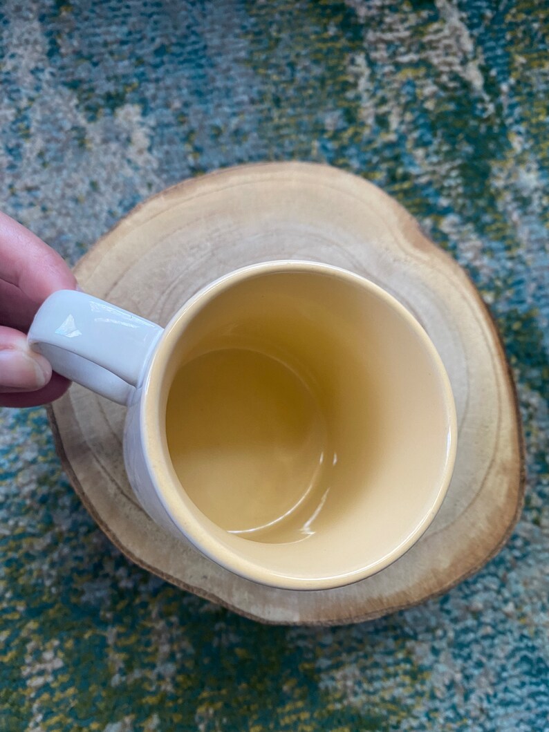 Może przedstawiać: Biały ceramiczny kubek z ż&oacute;łtym wnętrzem. Kubek jest pusty i stoi na drewnianej podstawce.