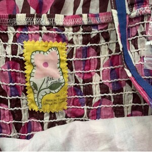 May include: A close-up of a pink and purple floral print fabric with a yellow Free People tag sewn on. The tag has a pink flower with a green stem and the text "Free People" in black. The tag also says "Size 10 Made in Taiwan fabrique en Taiwan".