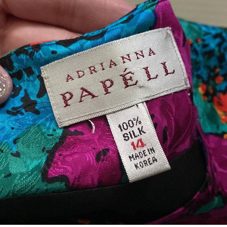 Puede incluir: Una etiqueta blanca con texto rojo que dice "Adrianna Papell" y una etiqueta blanca m&aacute;s peque&ntilde;a con texto negro que dice "100% Seda, 14, Hecho en Corea". La etiqueta est&aacute; sobre una tela colorida con un estampado floral.