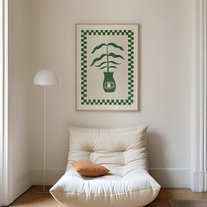 May include: A white, overstuffed armchair with a brown throw pillow sits in a room with white walls and wood floors. A green and white framed print of a plant in a vase hangs on the wall.