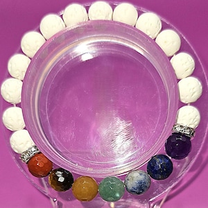 May include: A beaded bracelet featuring a mix of white, textured beads and colorful gemstone beads. The bracelet includes orange, brown, yellow, green, blue, and purple beads, with a silver accent. The bracelet is displayed on a clear, circular stand against a pink background.
