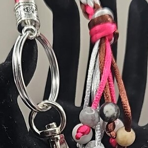 May include: A pink, brown, and white braided keychain with a silver key ring and a silver clasp. The keychain has a tassel with beads in various colors and patterns.