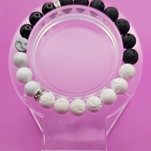 May include: A beaded bracelet featuring black lava rock beads, white textured beads, and a yin-yang bead. The bracelet includes silver-colored spacers. The bracelet is displayed on a clear, circular stand against a pink background.