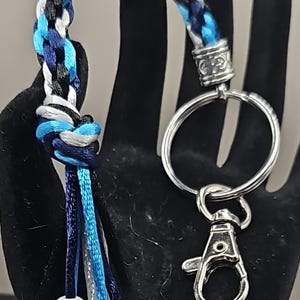 May include: A braided key chain with a silver ring and clasp. The braid is made of black, white, and blue cords. The keychain has a tassel with white and gray beads.