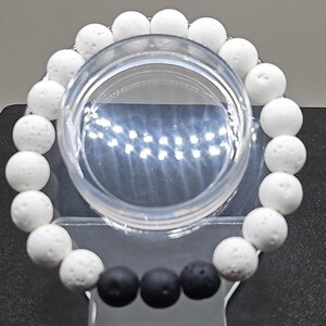 May include: A beaded bracelet featuring alternating white and black round beads. The white beads have a textured, porous appearance, while the black beads are smooth. The bracelet is displayed on a clear, circular stand, showcasing its design.