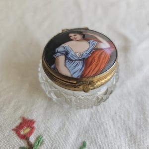 Può includere: Una piccola scatola rotonda in vetro trasparente con un coperchio a cerniera. Il coperchio è decorato con un'immagine dipinta di una donna con un vestito blu e uno scialle rosso.