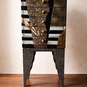 May include: A decorative chest of drawers with a unique design. The chest features a mix of black, white, and metallic colors, with gold-colored drawer pulls. The base has a textured, perforated metal design. The chest is on a wooden surface.