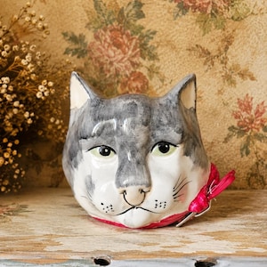May include: A ceramic cat head planter featuring a hand-painted grey and white coat with green eyes and a pink collar. The cat has visible whiskers, a small nose, and a smooth, glossy finish. The object is shaped as a feline head with pointed ears.