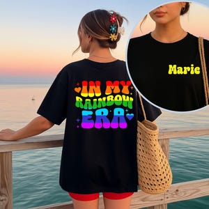 May include: Black t-shirt with the text "IN MY RAINBOW ERA" in rainbow colours on the back and "Marie" in yellow on the front. The shirt is worn with red shorts and a woven bag. The background shows a sunset over the ocean.