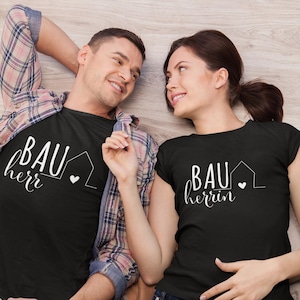 Puede incluir: Una pareja lleva camisetas negras a juego con texto blanco que dice "Bauherr" y "Bauherrin" con un pequeño icono de casa y un corazón.