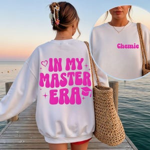 Könnte beinhalten: Weißes Sweatshirt mit dem Text "IN MY MASTER ERA" in leuchtendem Pink auf dem Rücken und dem Wort "Chemie" auf der Vorderseite. Das Sweatshirt hat einen Rundhalsausschnitt und lange Ärmel. Eine Abschlusskappe ist im Design enthalten.