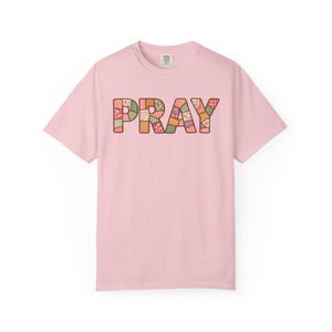 Puede incluir: Una camiseta de manga corta de color rosa claro con la palabra PRAY impresa en el pecho. Las letras están rellenas con un patrón de patchwork multicolor que consiste en varios diseños florales, de cuadros y geométricos en tonos tierra.