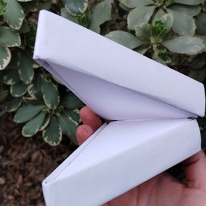 Puede incluir: Una caja blanca abierta, estilo origami, sostenida en una mano. La caja está hecha de papel plegado y tiene forma triangular. El fondo presenta hojas verdes y blancas y follaje marrón.
