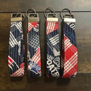 May include: Four fabric wristlet keychains featuring a patchwork design in red, white, and navy blue. The fabric displays football-themed graphics, including players and text, with silver-toned metal hardware and a circular split ring attached.