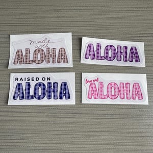 Puede incluir: Cuatro pegatinas rectangulares con la palabra "ALOHA" en un patrón de cuadros. Una pegatina dice "Made with ALOHA", otra dice "Raised on ALOHA", y una tercera dice "Love and ALOHA". Las pegatinas tienen un borde blanco.
