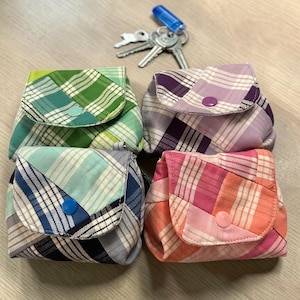 May include: Four small, square fabric pouches in green, purple, blue, and pink plaid patterns. Each pouch has a flap closure with a button. Keys with a blue key cover are placed above the pouches.