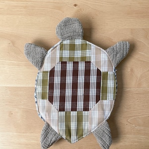 Op de afbeelding: Een schildpadvormig stoffen item met een centraal schild van patchwork ruitjesstof in tinten bruin, olijfgroen en beige. De kop en de vier flippers zijn gemaakt van een gestructureerde, lichtgrijze stof met een zachte uitstraling.