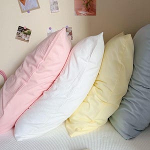 以下が含まれることがあります： ライトピンク、ホワイト、ペールイエロー、ソフトブルーの無地のパステルカラーの長方形の枕が4つ並んでいます。それぞれの枕は質感のある生地の表面を持ち、壁に立てかけられています。