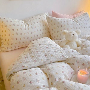 May include: A white bed with a floral patterned duvet cover and matching pillowcases. A white stuffed animal sits on the bed, and a pink candle is on a small tray.