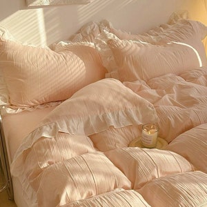 May include: A soft, peach-colored bedding set with ruffled edges. The set includes a duvet cover, two pillowcases, and a decorative throw pillow. The bedding is arranged on a bed with a white headboard and a white rug.