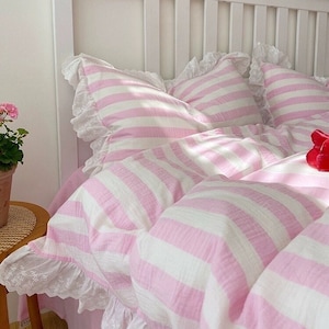 May include: A white bed with a pink and white striped bedding set. The bedding has a white lace trim. The headboard is white wood with vertical slats.
